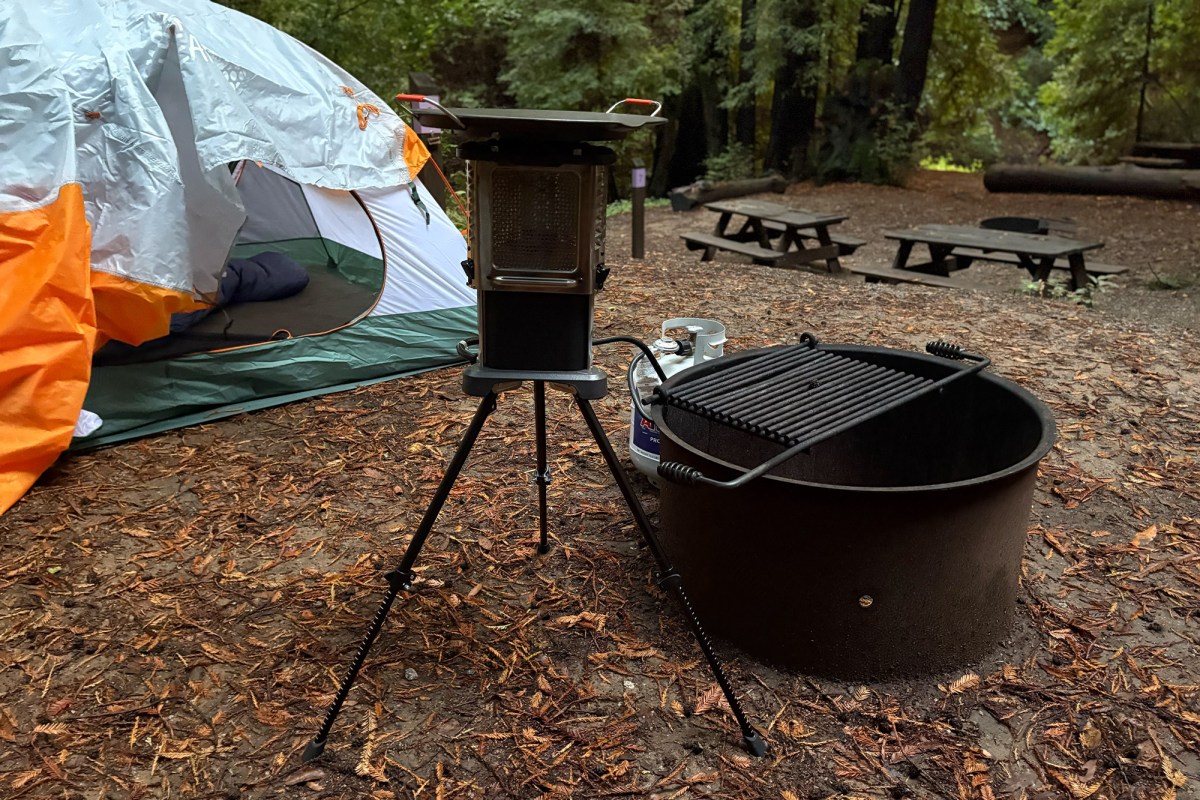 Ignik Skullet setup on tripod legs at a forest campsite next to a tent and a fire ring.