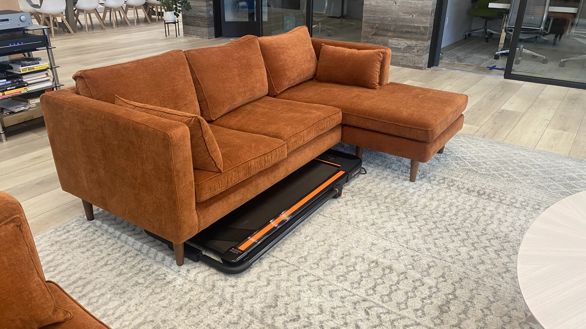 A slim UREVO treadmill being slid horizontally underneath a grey living room couch.