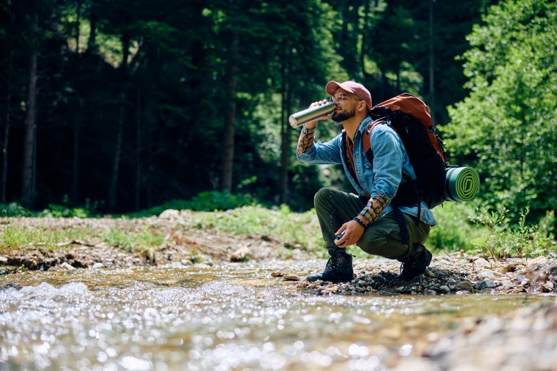 A hiker tiltng their head back to drink water from a bottle in a sunny forest.
