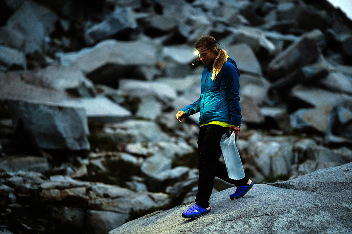 A woman walking across a wet rock near a campsite wearing blue ThermoBall Traction Mules.