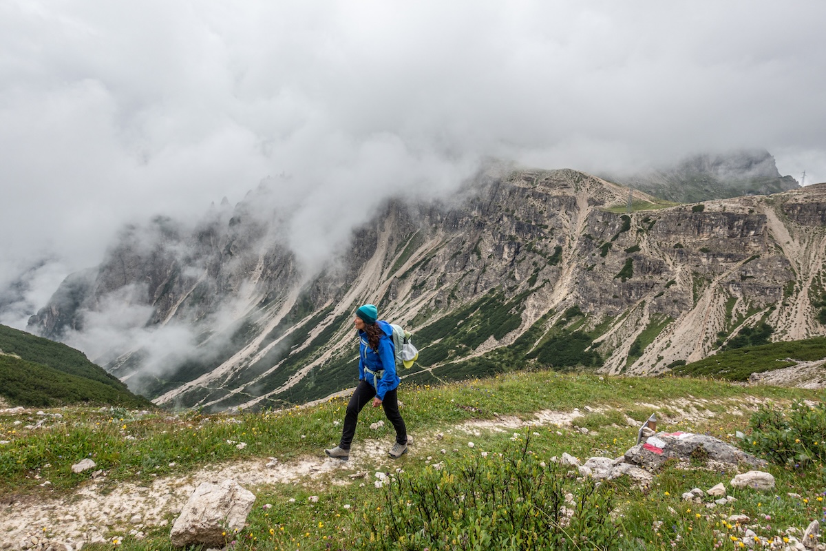 A woman hiking in a blue hardshell jacket with mountains in the background.