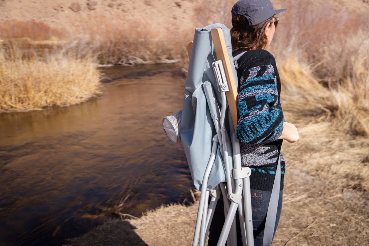 A person carrying a folded camping chair over their shoulder using integrated straps.