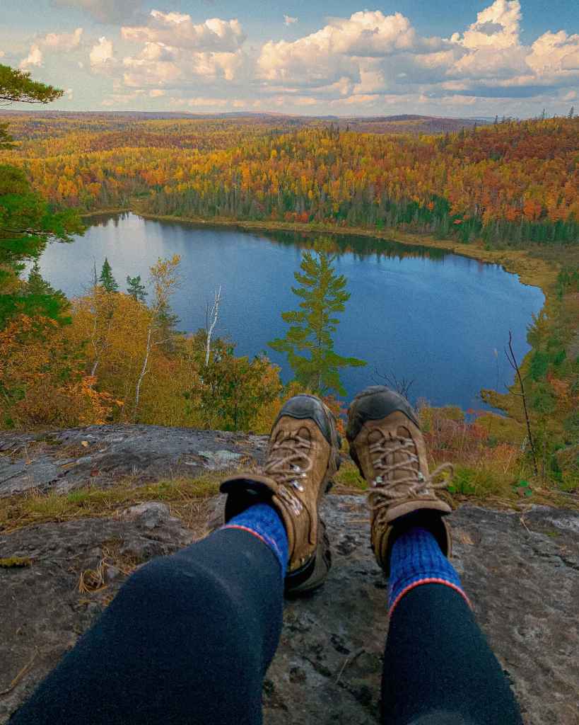 Hiking boots dangling above a clear blue lake with vibrant autumn forest colors in the background.