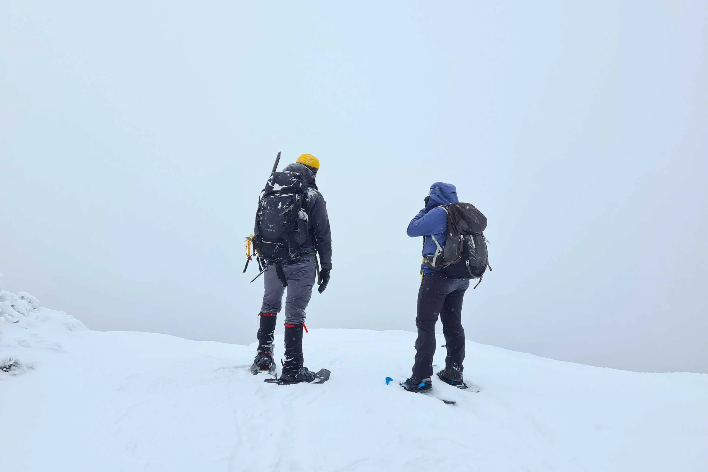 Two hikers traversing a snowy, wind-swept mountain ridge in the Adirondacks wearing full-length gaiters.