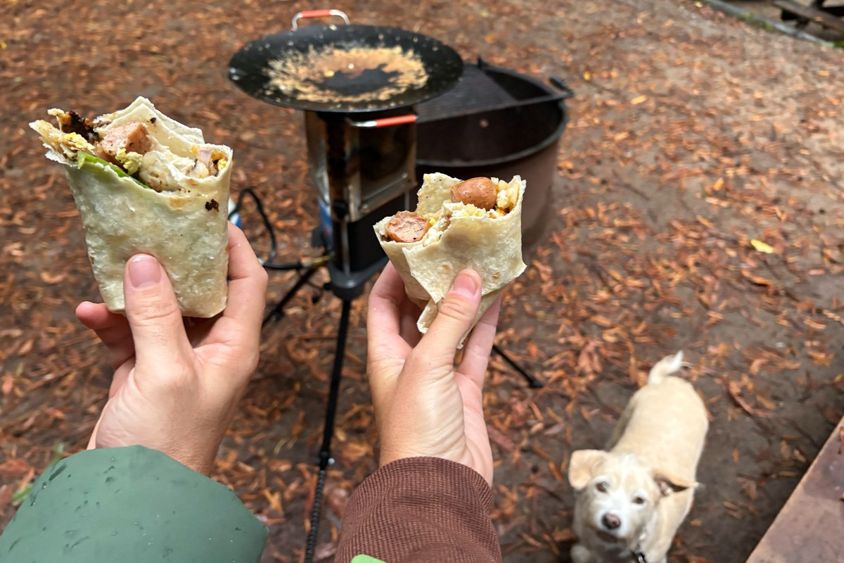 Two hands holding breakfast burritos at a campsite with the Ignik Skullet and a dog in the background.