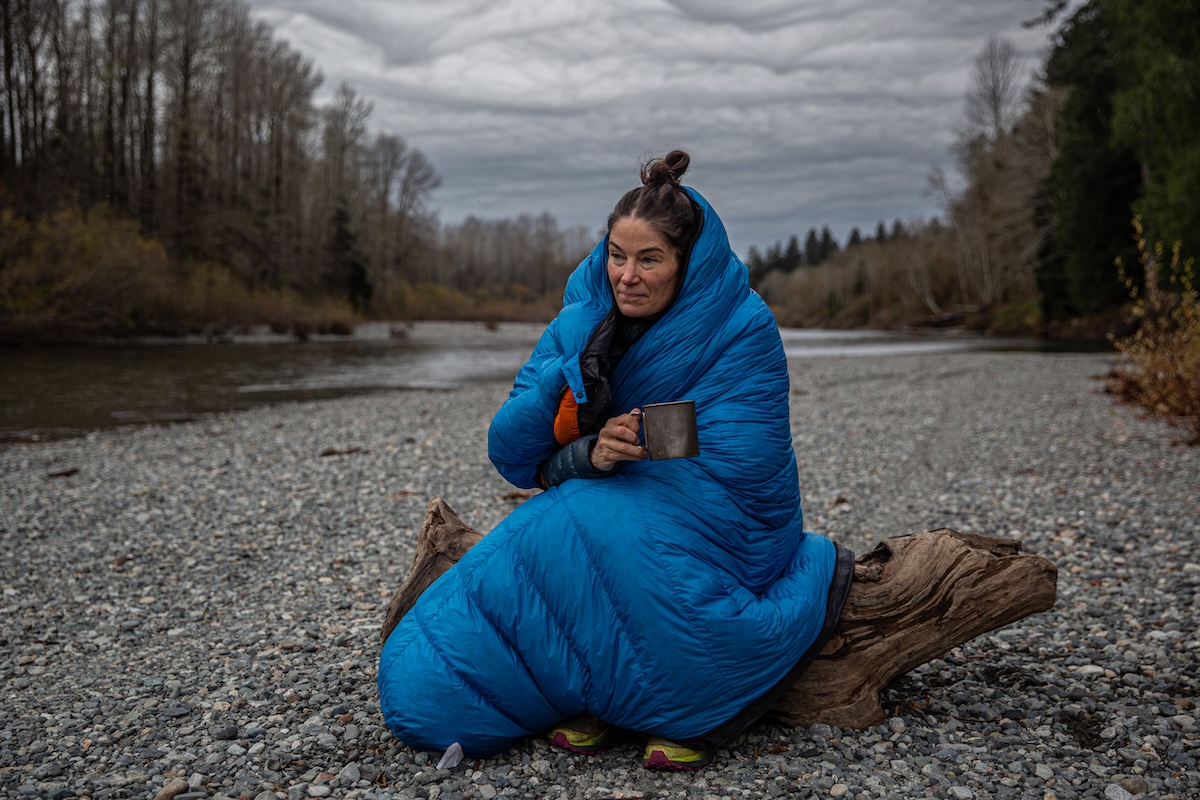 A woman sits on a log wrapped in a blue Feathered Friends Hummingbird UL sleeping bag.