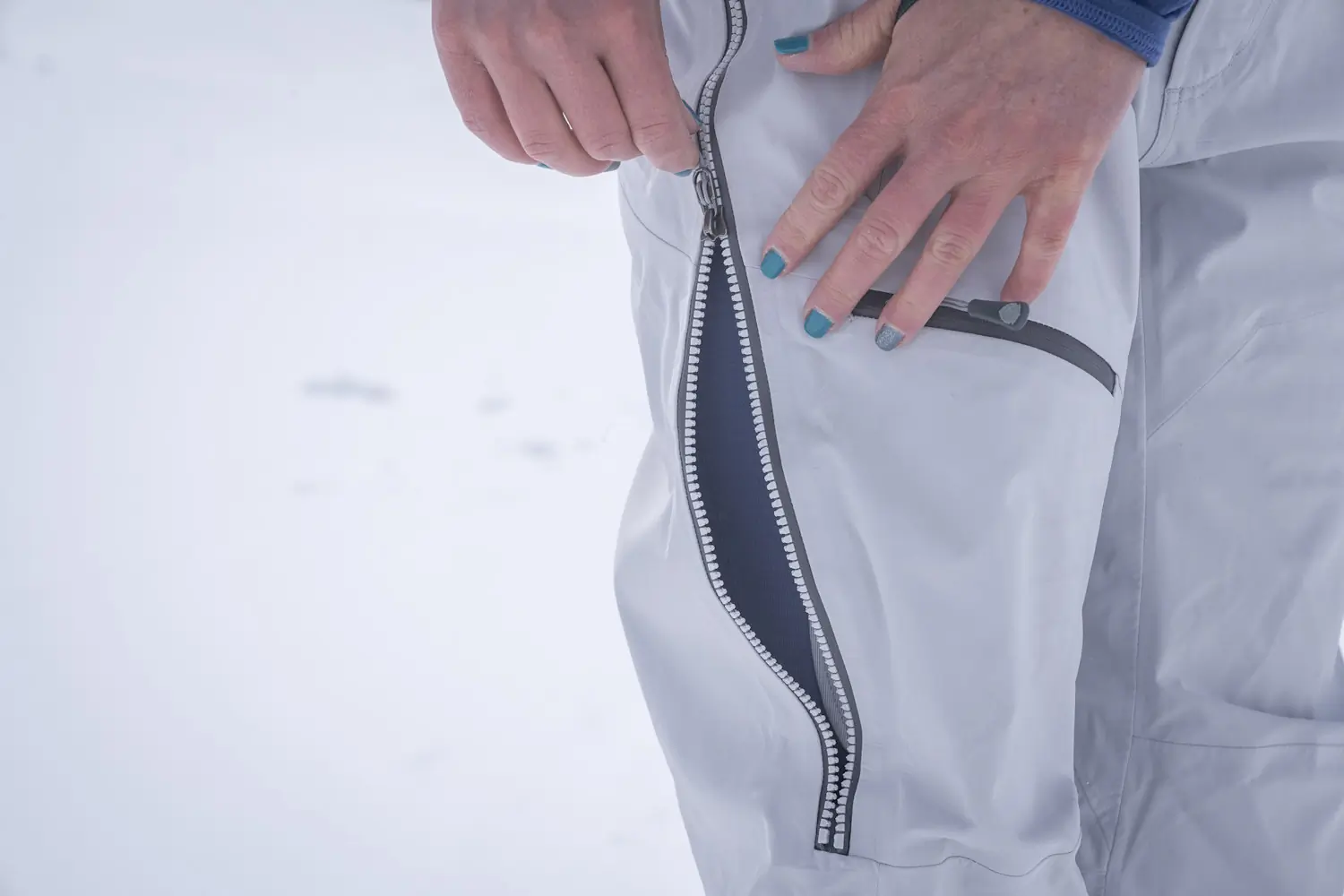 A close-up view of a person reaching for the side ventilation zipper on technical ski bibs.
