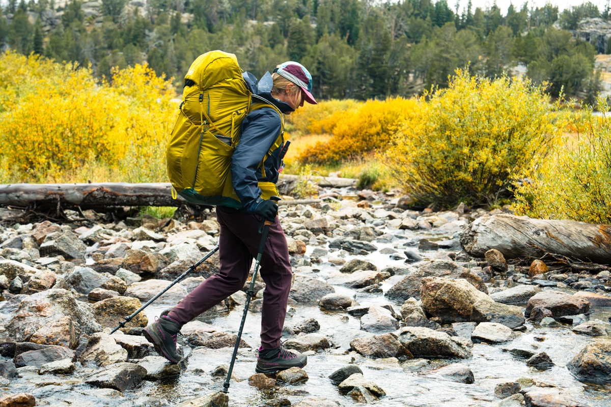 A hiker wearing waterproof boots walks across a shallow, clear mountain stream surrounded by yellow fall foliage.