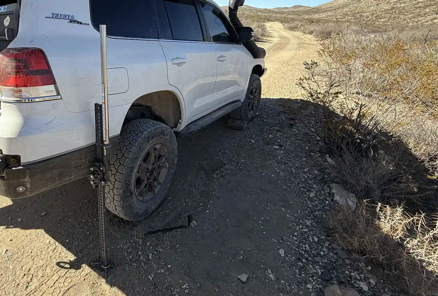 A Hi-Lift Jack being used to elevate a modified off-road vehicle in a sandy desert environment.