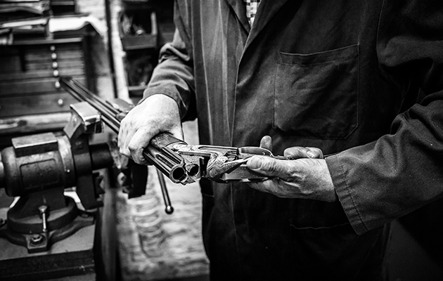 A gunsmith working on a shotgun in a workshop surrounded by precision tools.