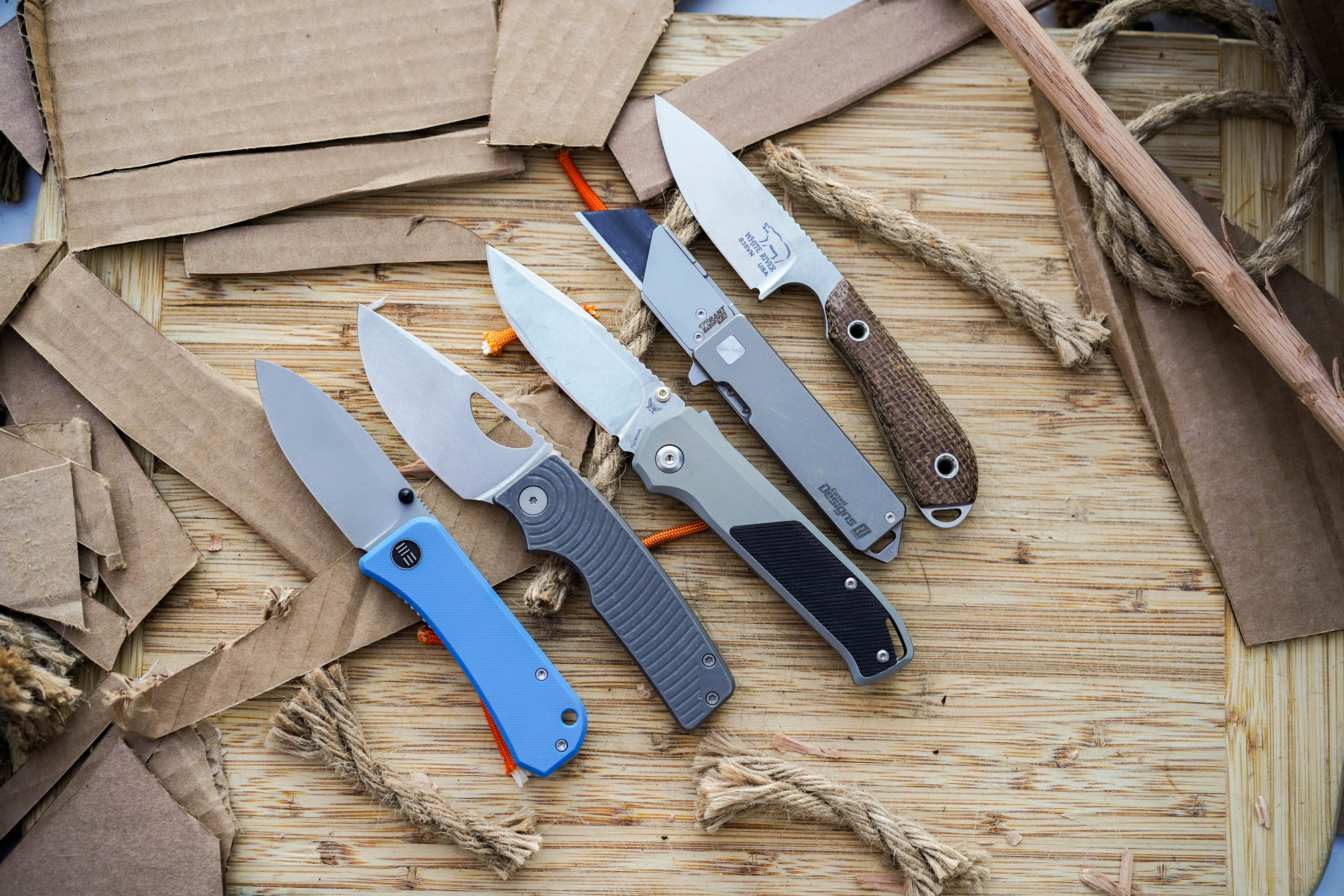 A large selection of pocket knives laid out on a wooden table for comparative testing.
