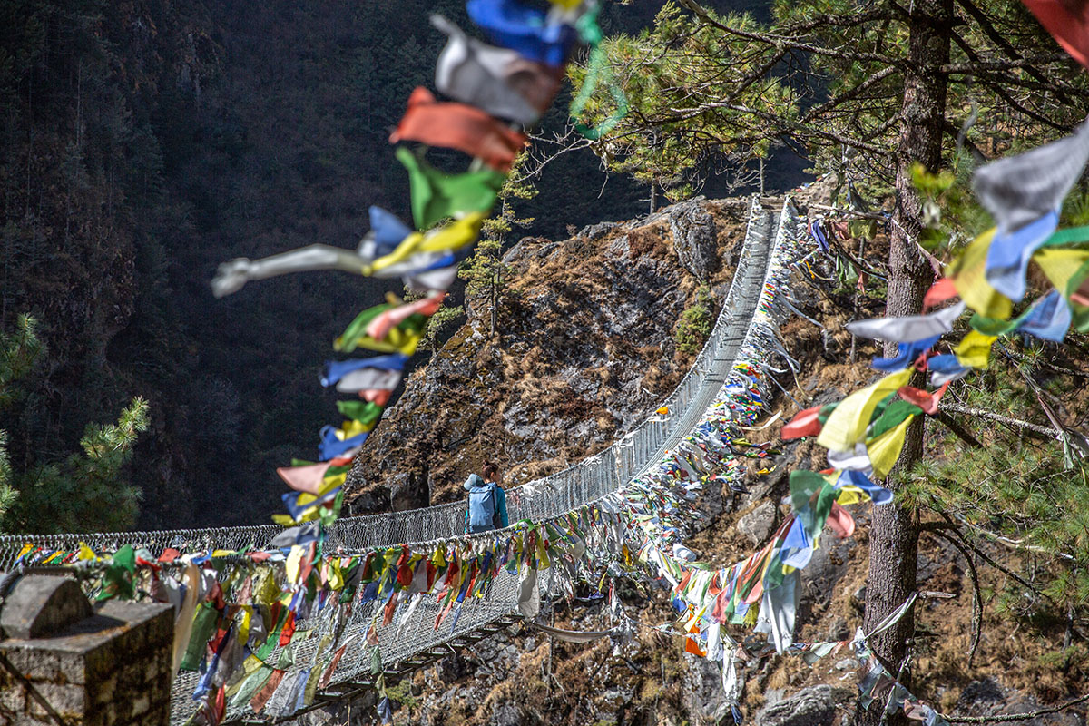 Trekkers crossing a high suspension bridge draped with prayer flags leading toward Namche Bazaar.