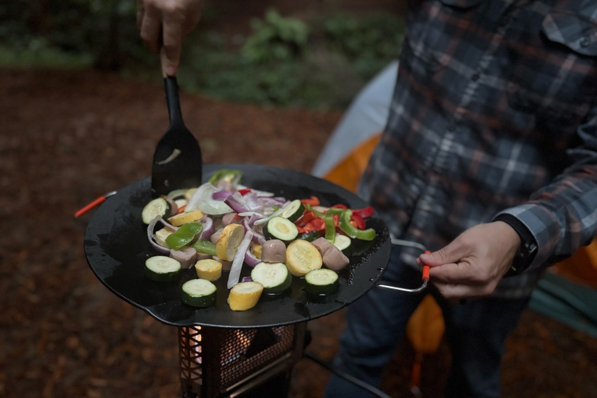 Person stirring vegetables and sausage in a skillet on top of the Ignik Skullet stove.