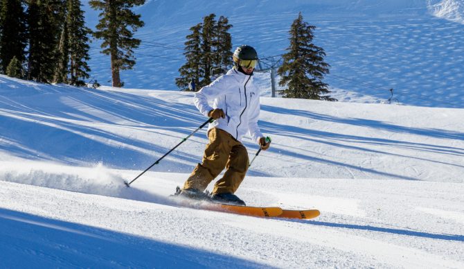 A skier in a blue Helly Hansen Alpha 4.0 jacket carving through deep powder snow.