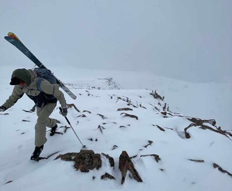 A backcountry skier hiking a rocky snowy ridge with skis strapped to their pack.