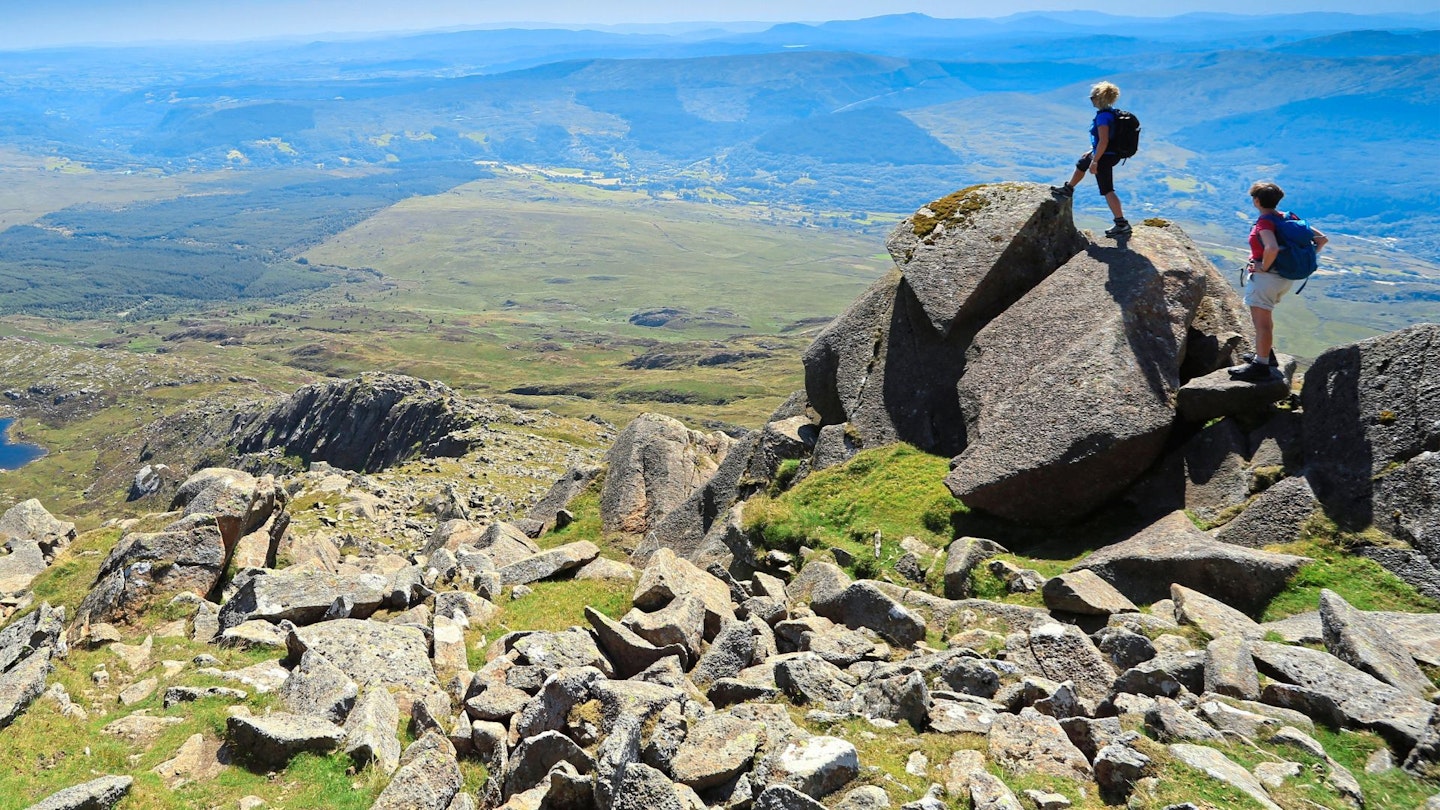 Two hikers walking through a sunlit mountain trail wearing lightweight daypacks.
