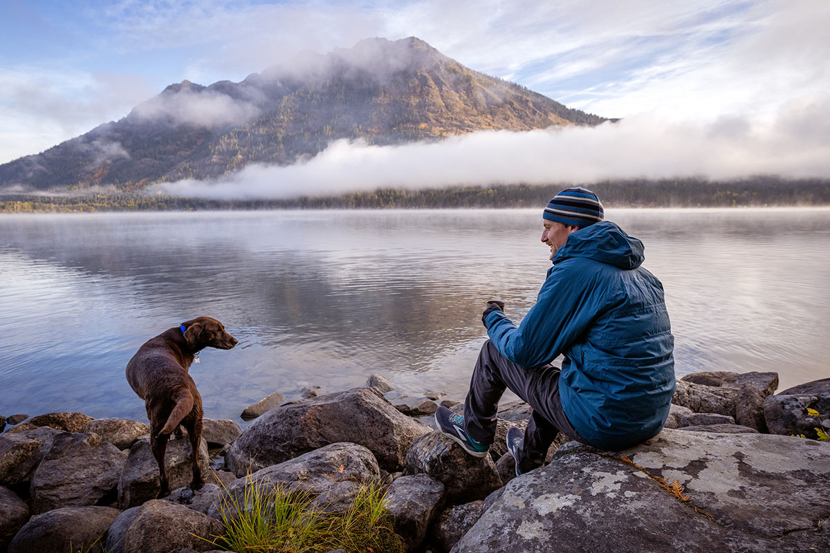 A person wearing the REI Stormhenge jacket sitting by a foggy mountain lake.