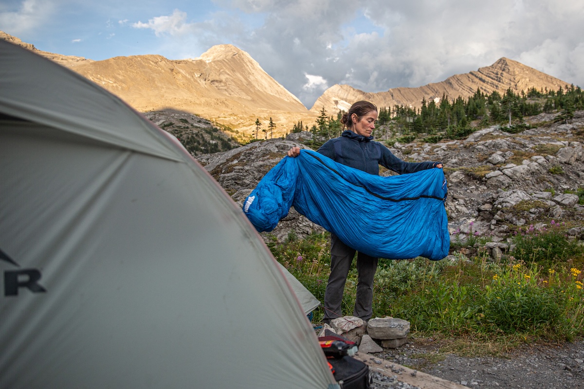 A woman holds a Feathered Friends Hummingbird UL sleeping bag outside a tent.
