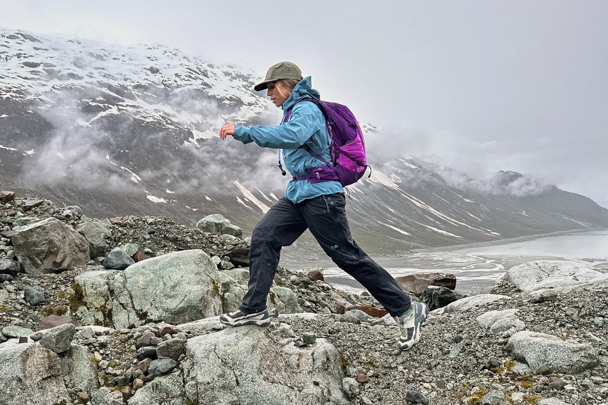 A woman wearing Salomon hiking shoes hops between large boulders in a misty mountain setting.