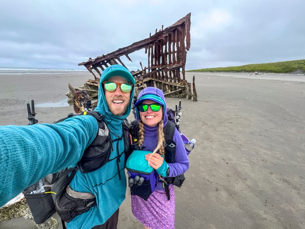Thru-hikers posing in front of the rusted Peter Iredale shipwreck on a sandy beach.