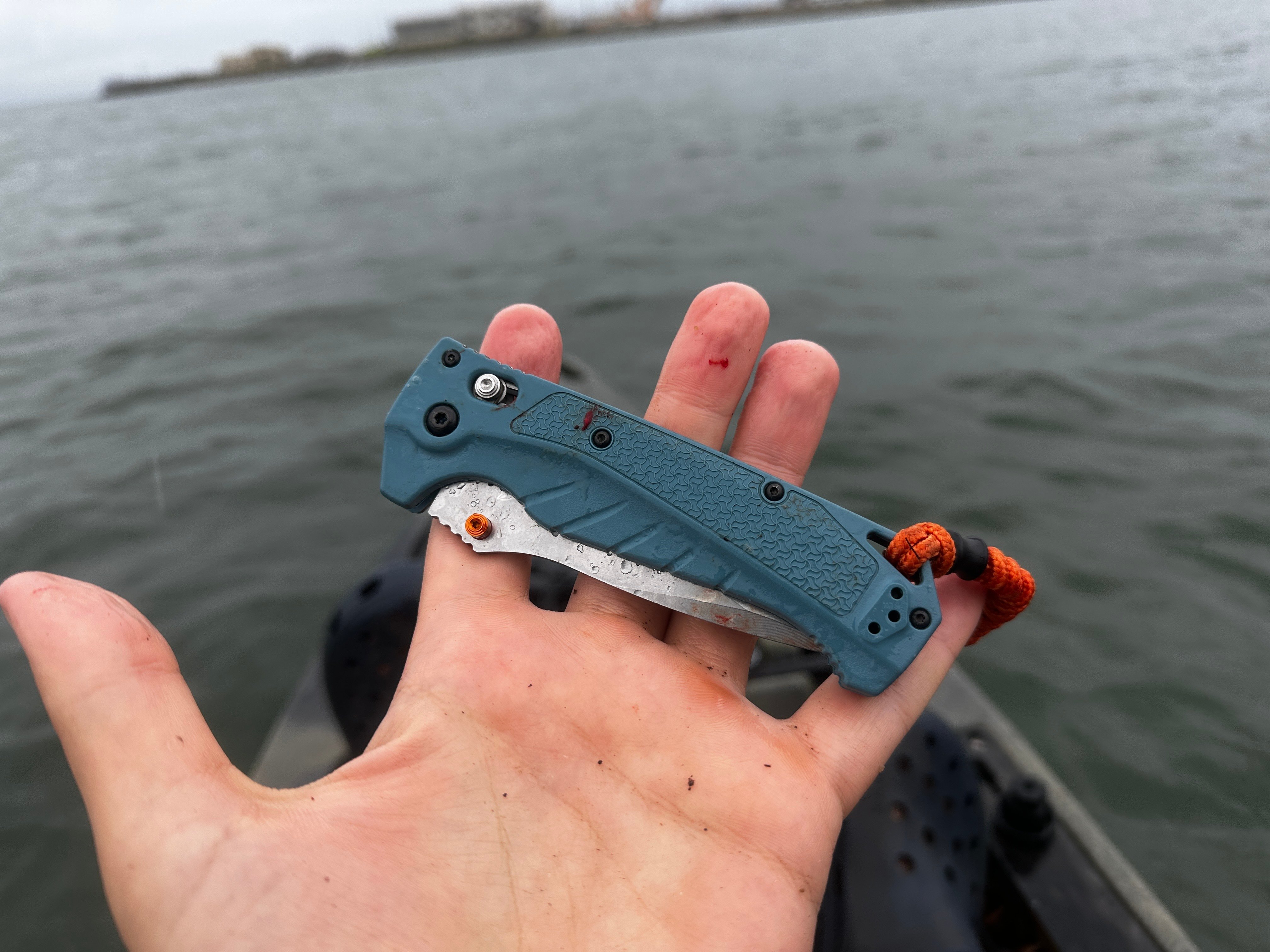A blue folding knife held by a person with a kayak and water in the background.