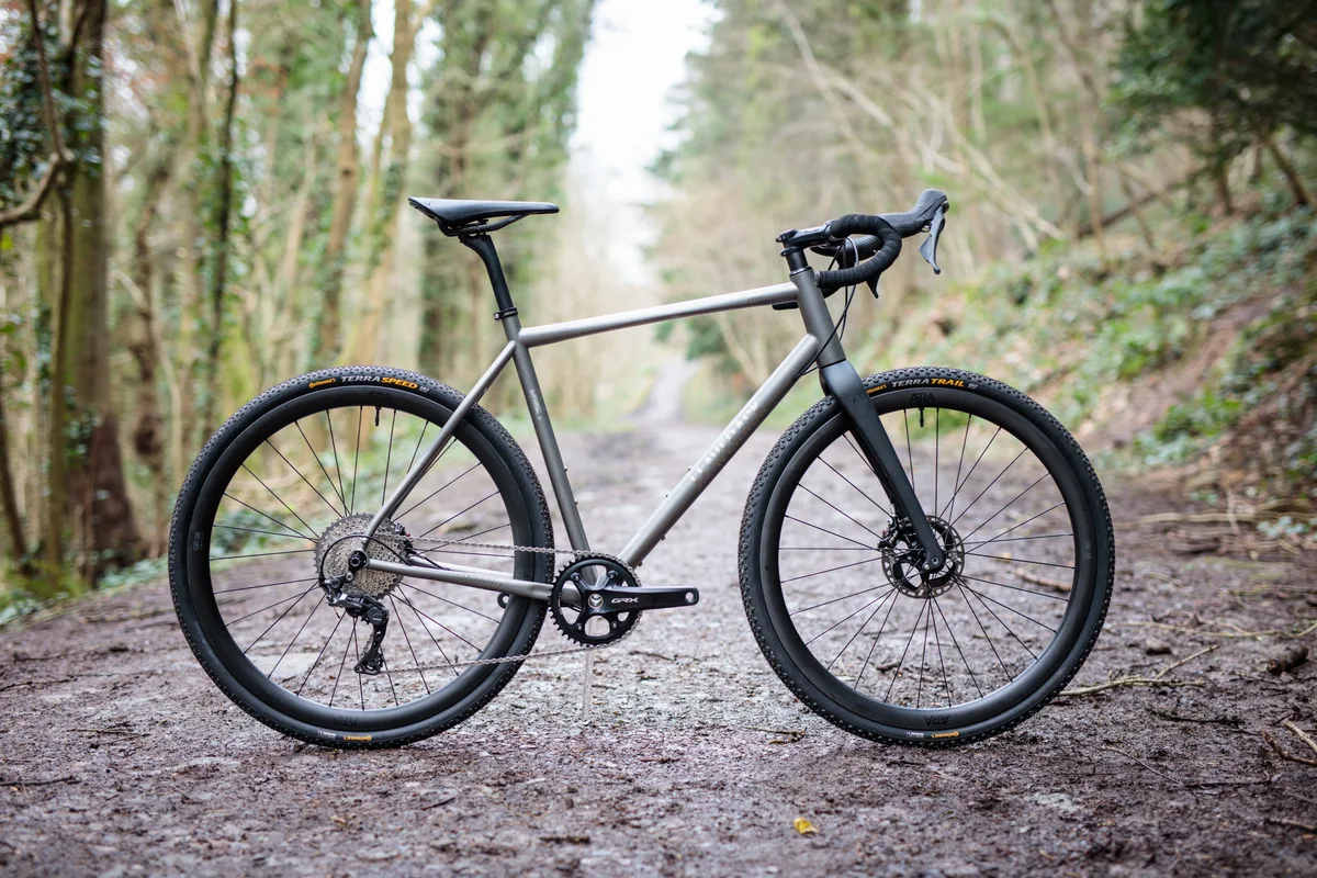 A cyclist riding a titanium gravel bike on a wooded dirt track.