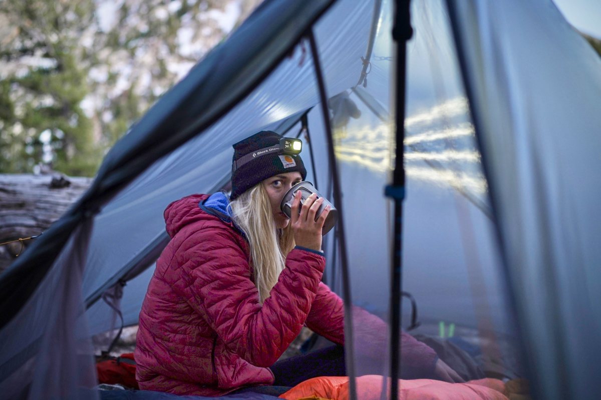 A backpacker sitting inside a tent with a warm drink, wearing a quilt-style synthetic jacket.
