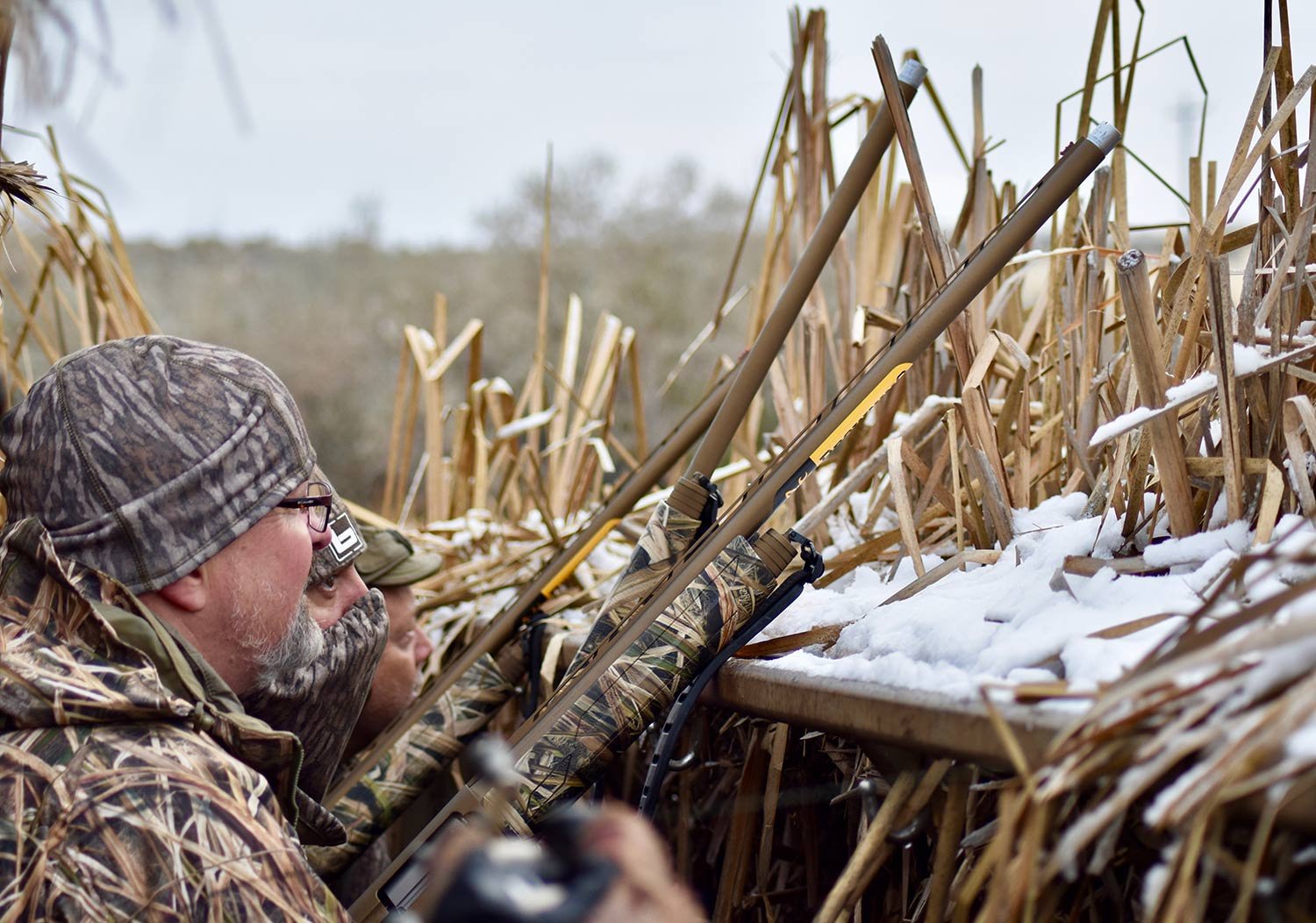 A hunter wearing camouflage aiming a shotgun from a wooden hunting blind.