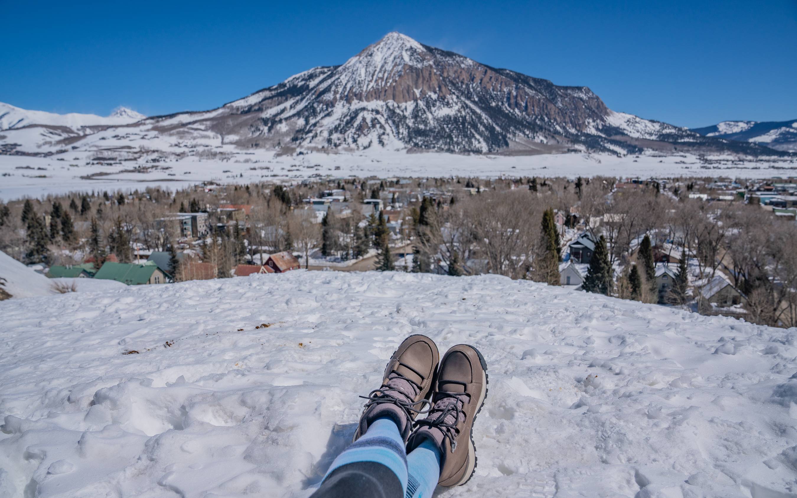 A pair of winter boots standing in deep snow with the Crested Butte mountain peaks in the distance.