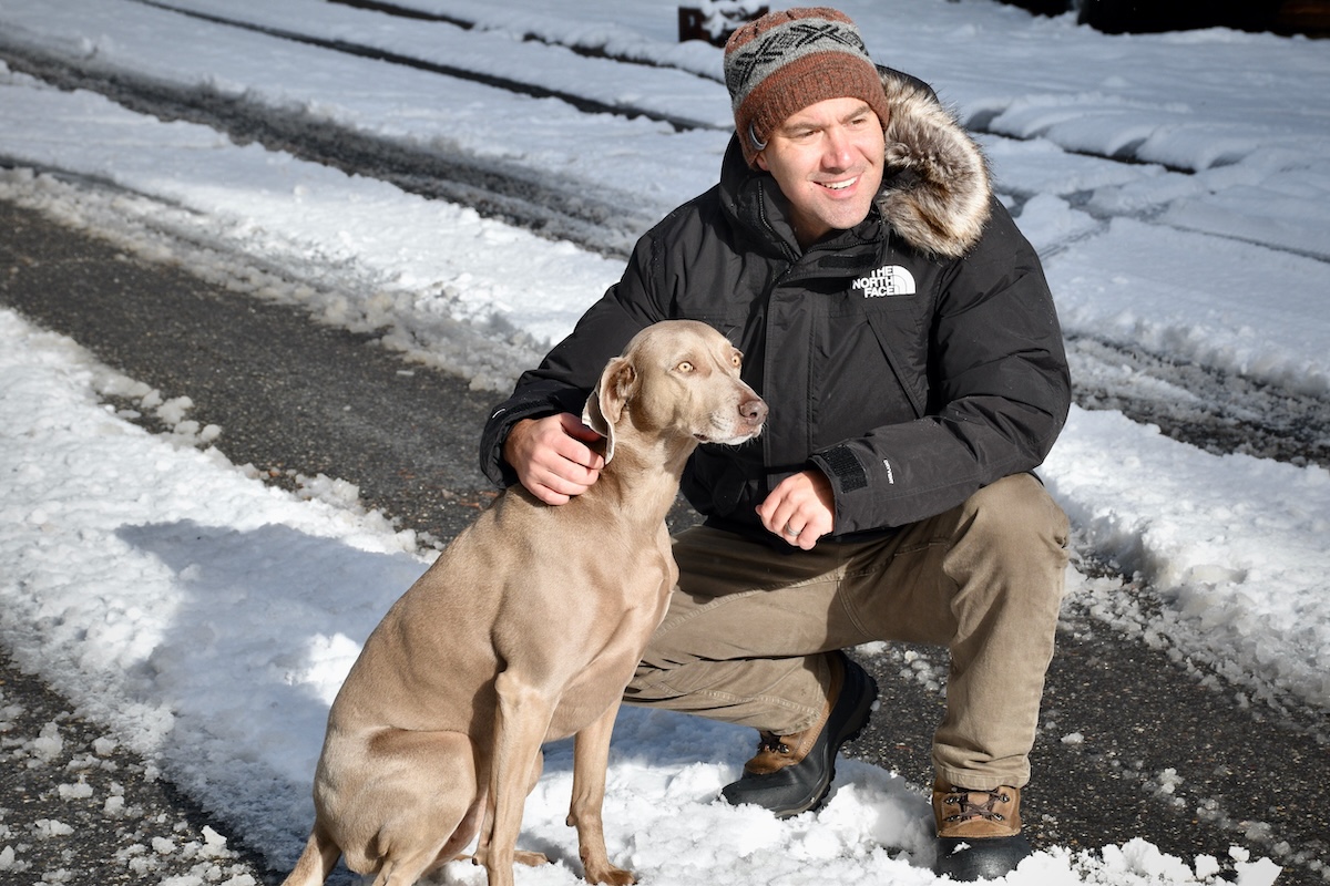 A man wearing a black North Face McMurdo parka kneeling with a dog on a snowy road.