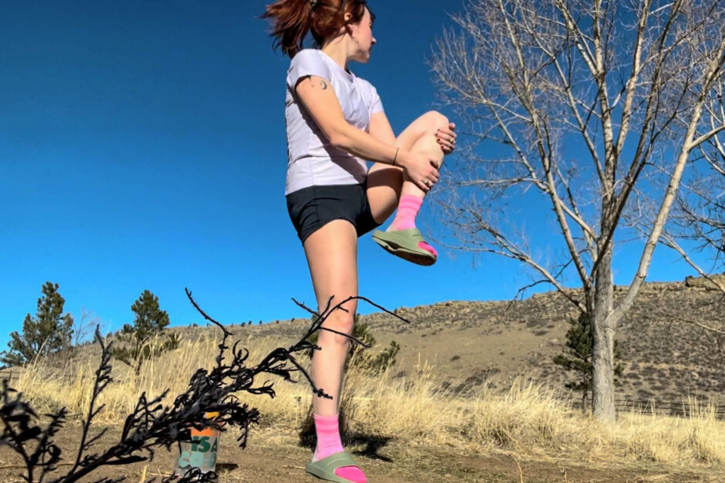 A runner sitting on a trail edge wearing Oofos OOahh recovery slides after a run.